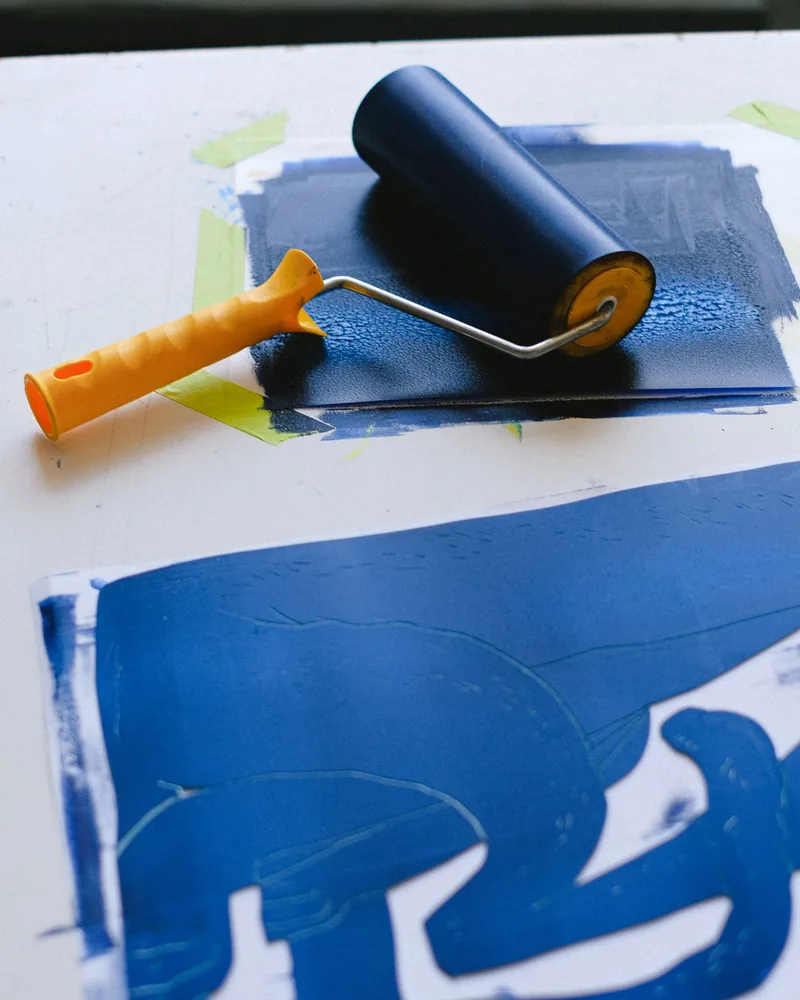 Rodillo de entintar cargado de tinta azul sobre la placa de entintado, junto a una estampa de linóleo recién tirada
